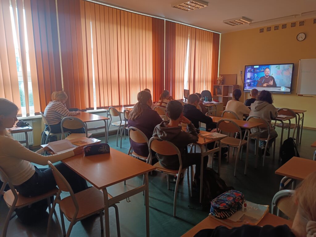 Klasa szkolna z uczniami siedzącymi tyłem do obiektywu, obserwującymi film edukacyjny na dużym monitorze. Na ekranie widać mężczyznę (prawdopodobnie prowadzącego) przemawiającego w filmie. Pomieszczenie jest dobrze oświetlone przez duże okna z pionowymi żaluzjami.