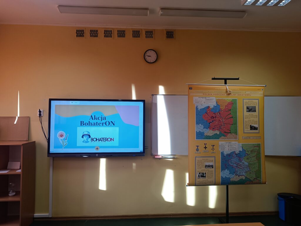 Zdjęcie klasy szkolnej z widoczną częścią ściany. Na środku powieszony jest duży monitor, na którym wyświetla się ekran powitalny akcji "BohaterON - Włącz Historię" z logo. Obok monitora stoi tablica suchościeralna i rozwinięty plakat dydaktyczny "Polska podczas II Wojny Światowej".
