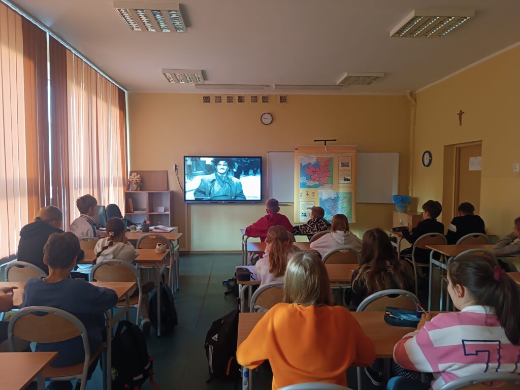 Klasa szkolna z uczniami siedzącymi przy ławkach i oglądającymi film na dużym monitorze. Na ekranie widać czarno-białe zdjęcie historyczne przedstawiające młodego żołnierza z okresu II Wojny Światowej. Na ścianie, obok monitora, wisi plakat "Polska podczas II Wojny Światowej".